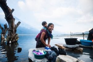 Woman carrying a child on her back and washing clothes by a lake